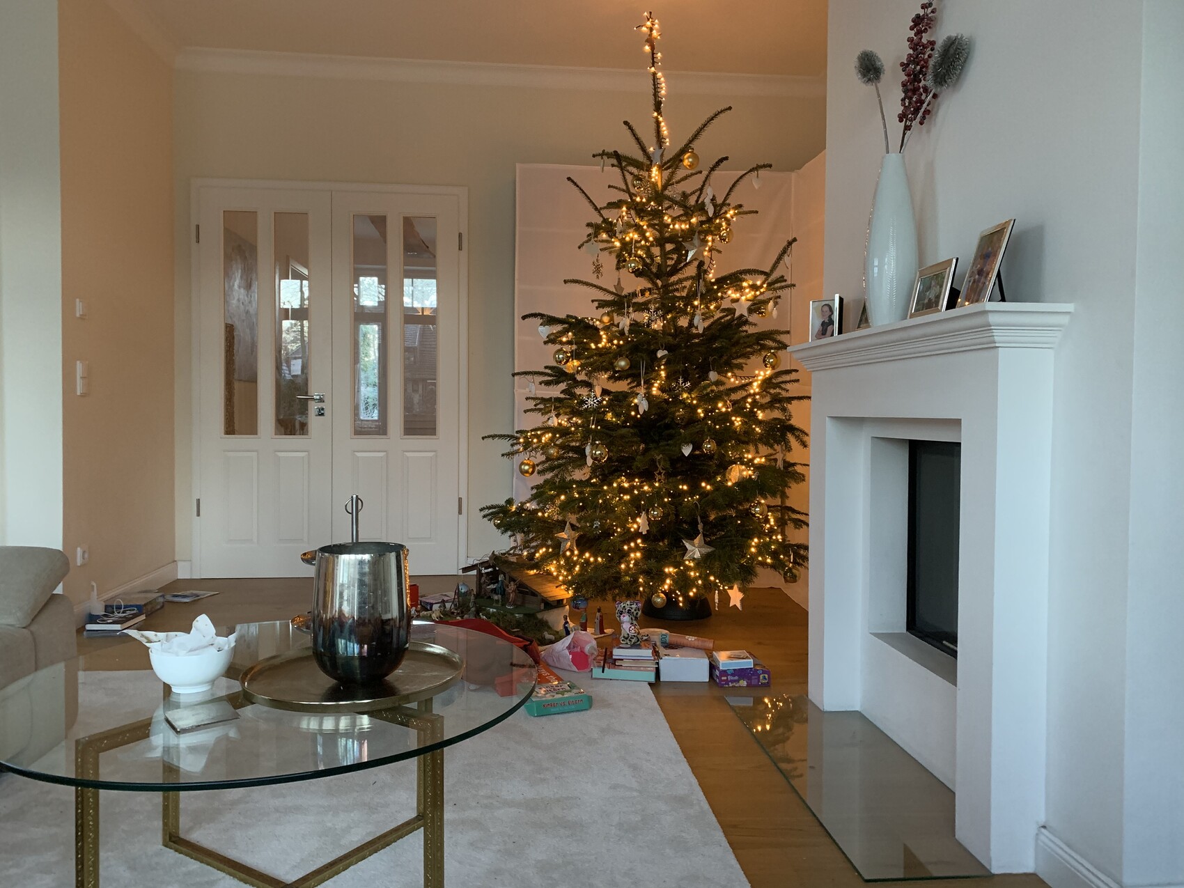 Wohnzimmer mit festlich geschmücktem Weihnachtsbaum, Geschenken darunter, Kamin und Glas-Couchtisch.