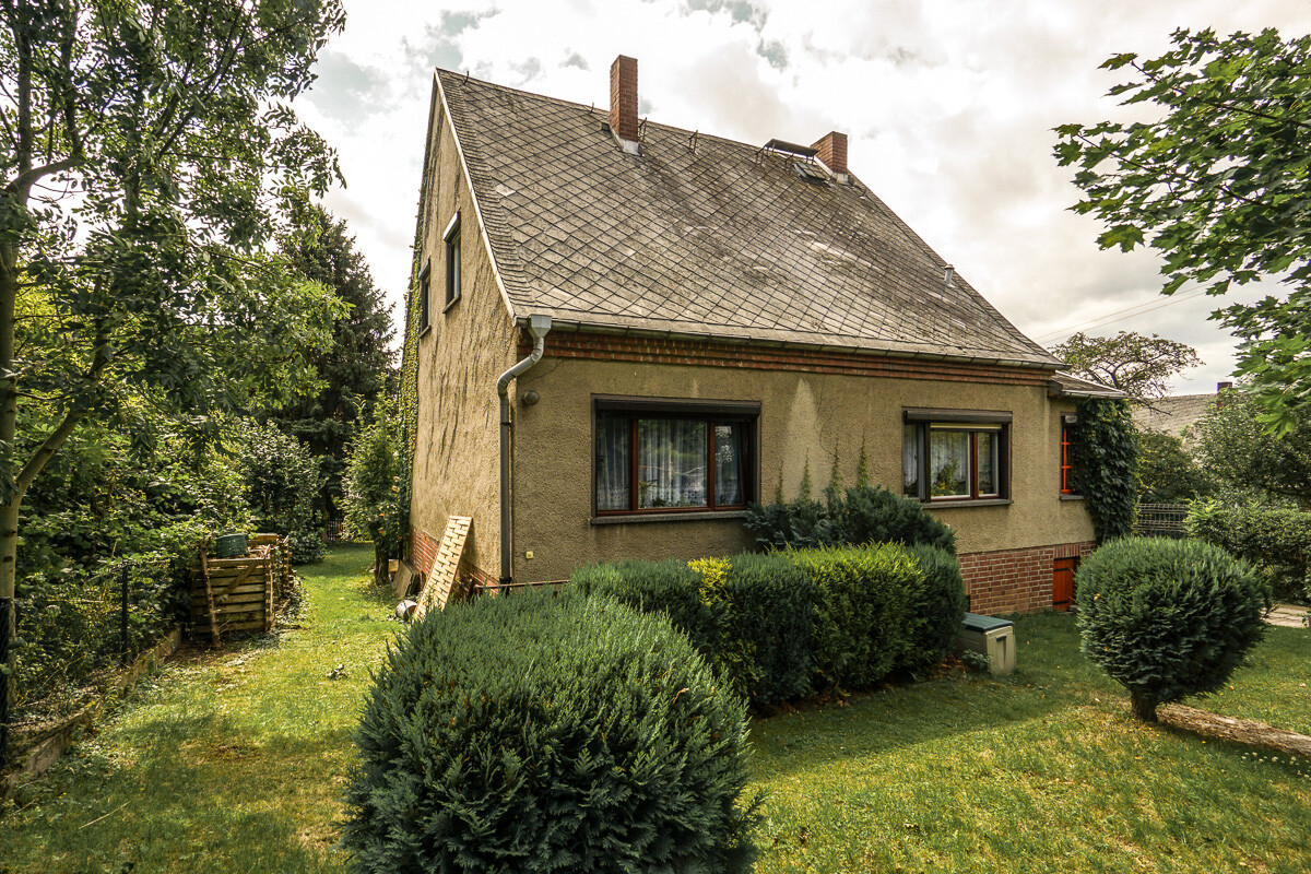 Kleines Einfamilienhaus mit steilem Dach, Garten und Sträuchern im Vordergrund.