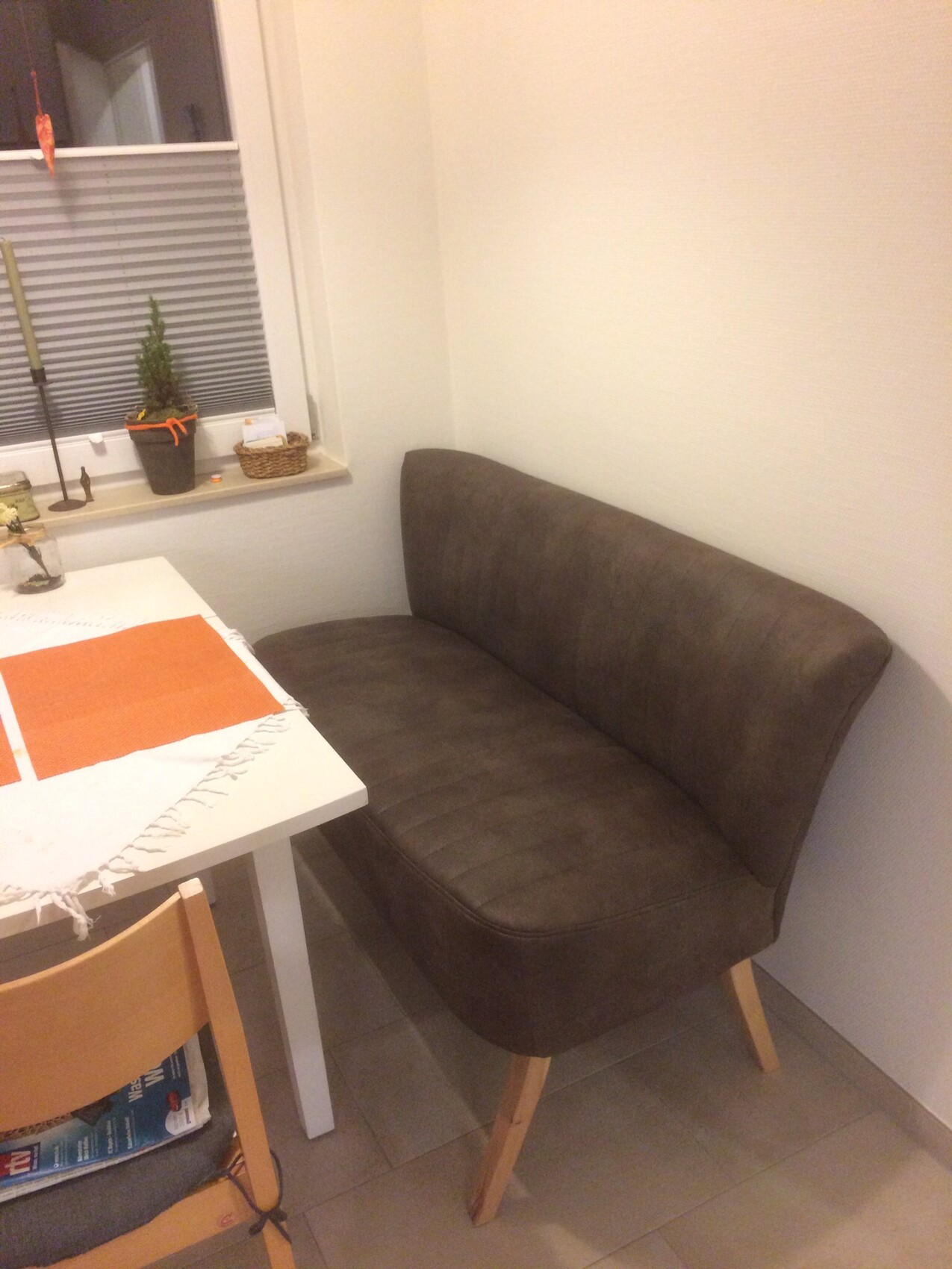 Kleines Wohnzimmer mit braunem Stoffsofa, weißem Tisch, orangenen Platzdeckchen und Fensterjalousien.