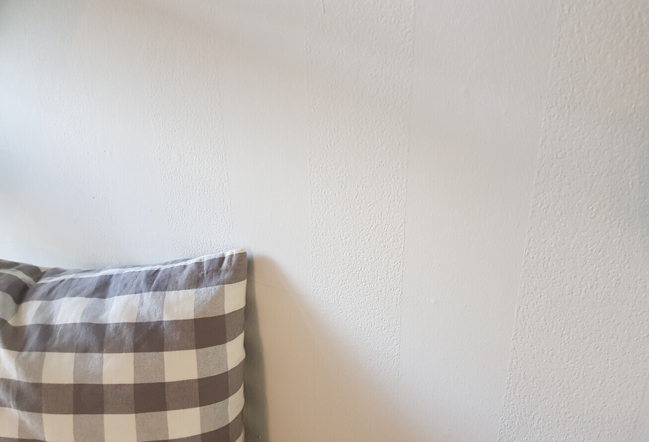 Weiße, texturierte Wand; links unten liegt ein graues kariertes Kissen.