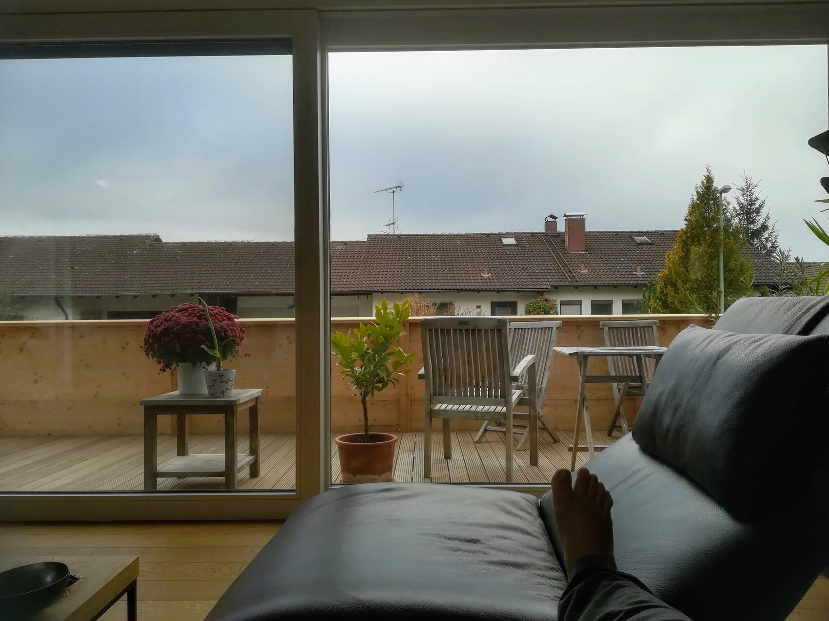 Wohnzimmer mit Balkonblick: schwarze Ledercouch im Vordergrund, Fuß sichtbar, Tischgruppe und Pflanzen.