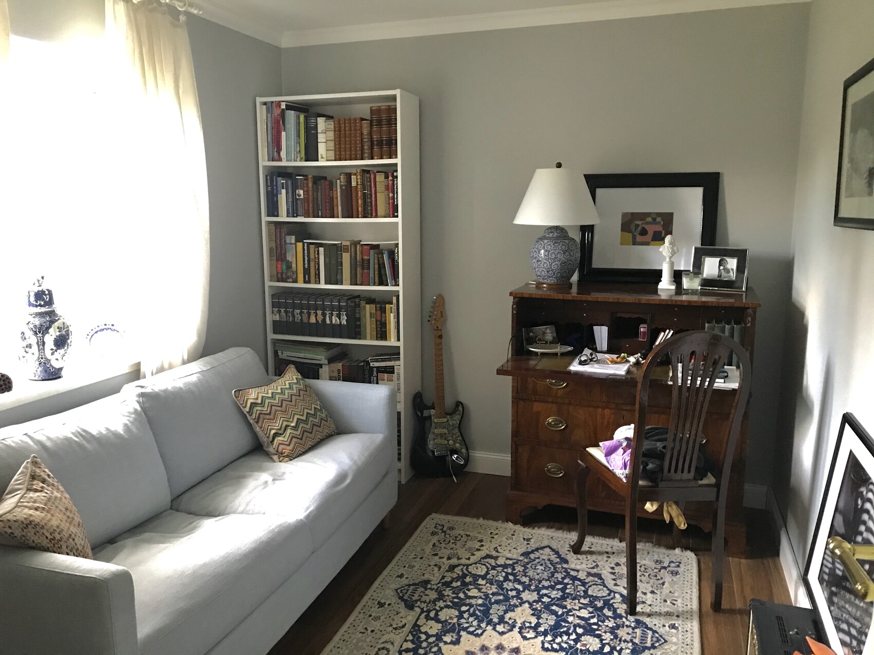 Helles Wohnzimmer: weiße Couch neben Bücherregal, E-Bass, Holzschreibtisch mit Lampe und Deko.