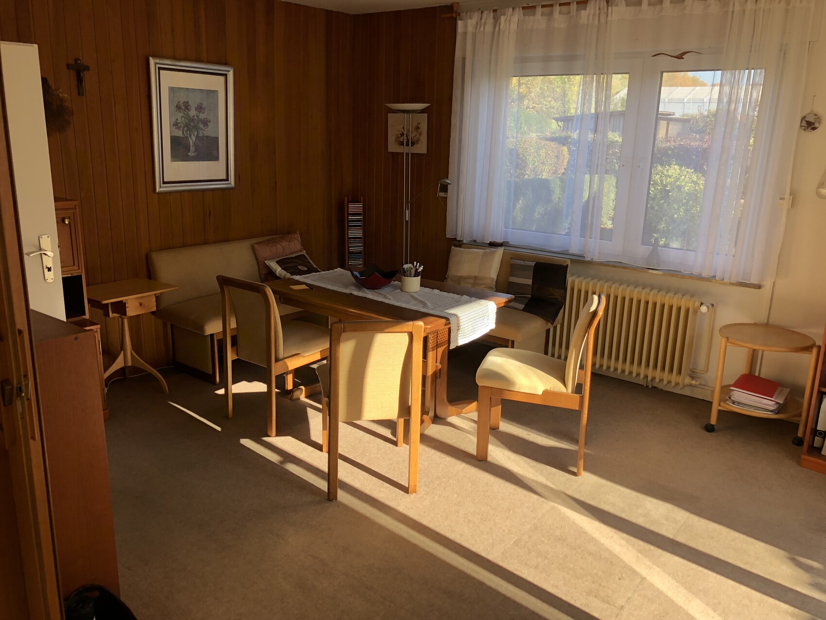 Wohnzimmer mit Esstisch, Stühlen, Sofa, Holzvertäfelung und Fenstern mit weißen Vorhängen.