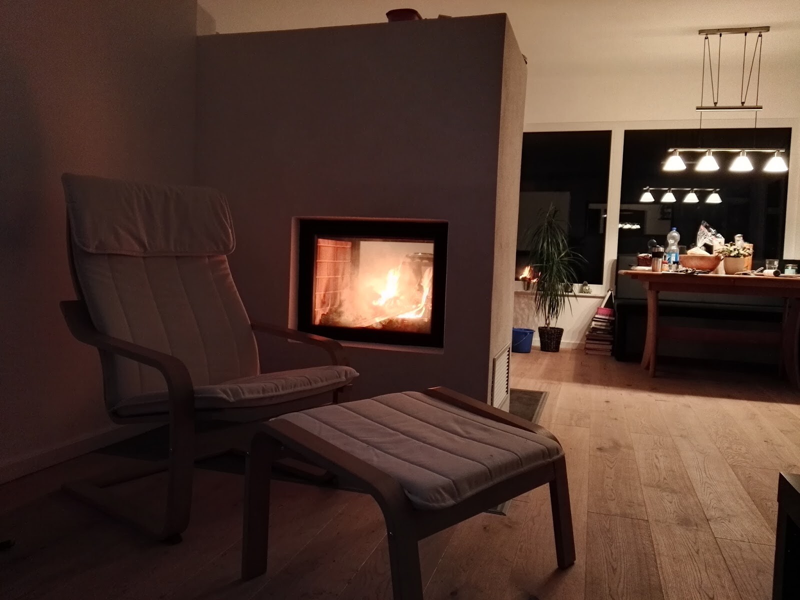 Wohnzimmer mit brennendem Kamin, Lounge-Sessel und Hocker im Vordergrund, Esstisch im Hintergrund.