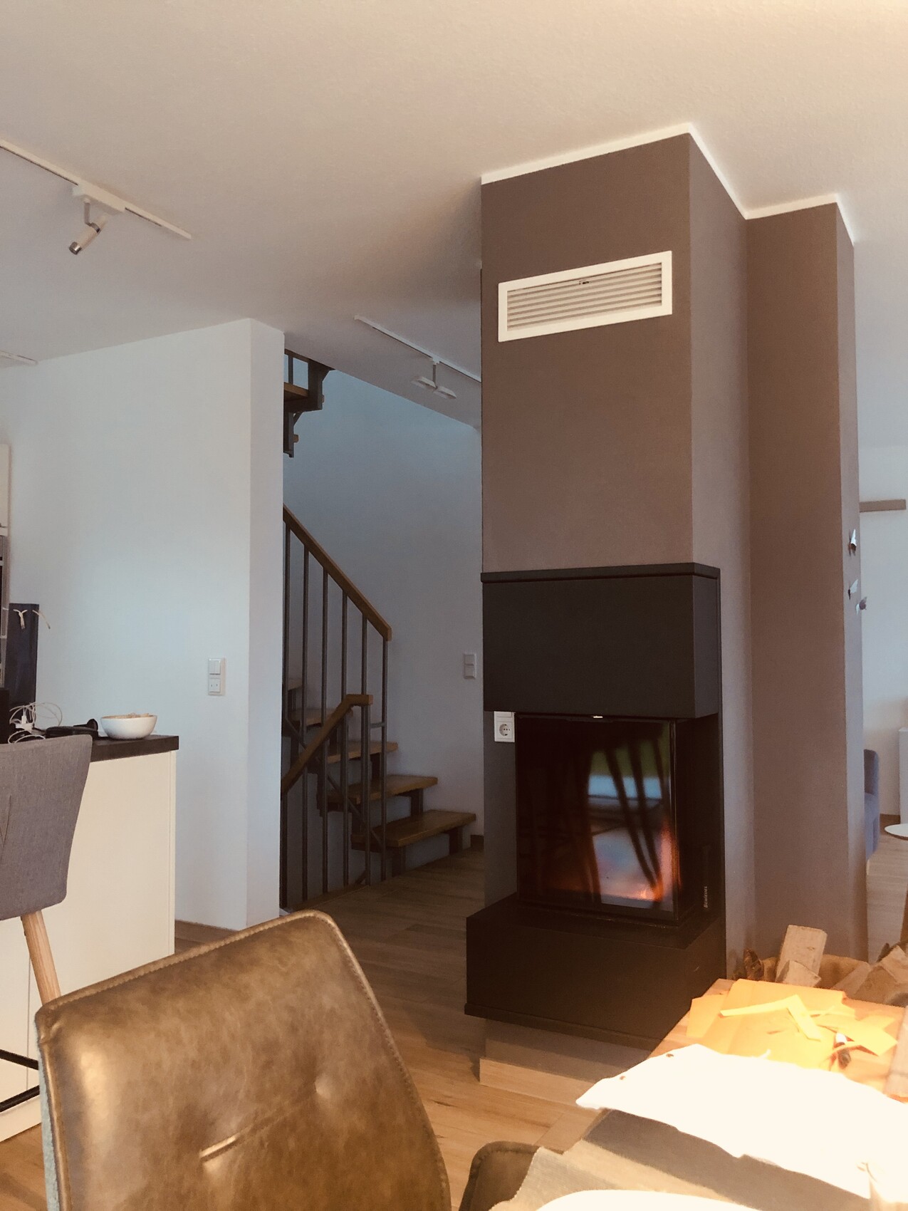 Wohnzimmer mit großer grauer Kamin-Säule, Feuer im Ofen, Treppe links, Möbel im Vordergrund.