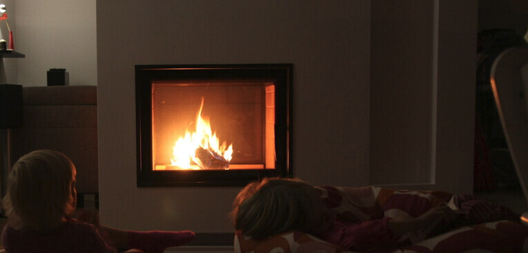 Wohnzimmer mit brennendem Kamin; links sitzt ein Kind, vorne liegt eine Person unter Decke