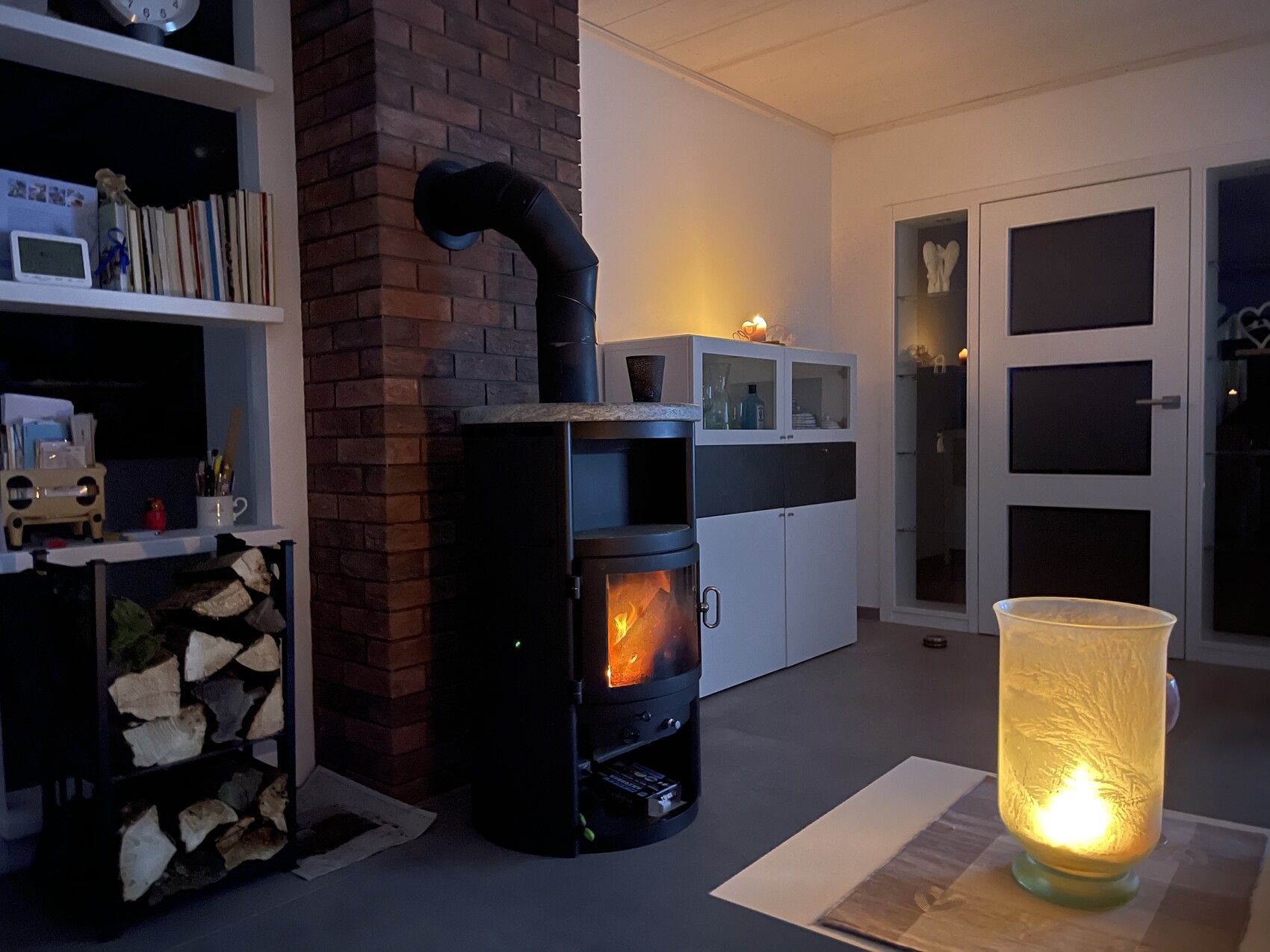 Wohnzimmer mit brennendem Kaminofen, Holzstapel, Bücherregal und warm leuchtender Tischlaterne.