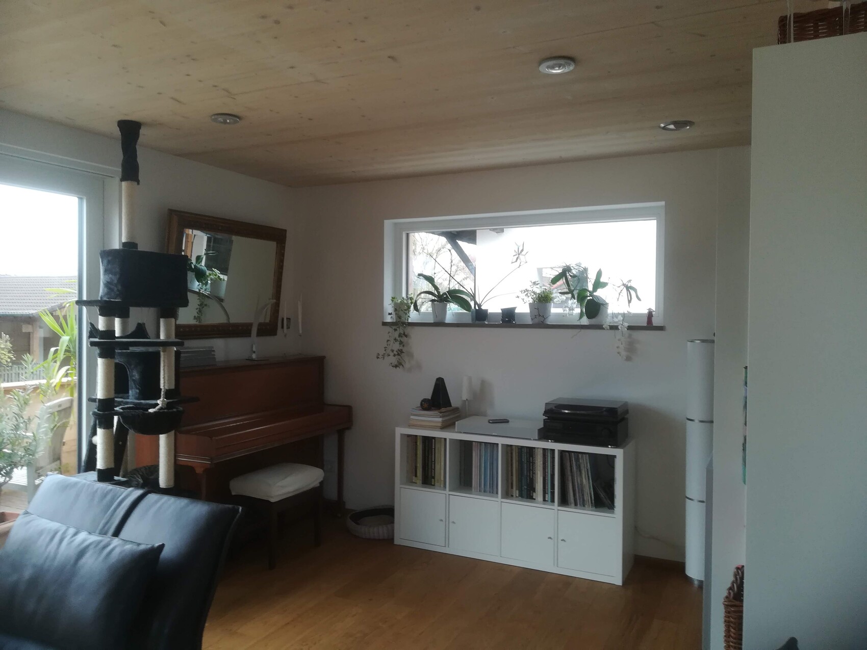 Wohnzimmer mit Klavier, Katzenbaum, weißem Schrank voller Schallplatten und Fensterpflanzen.