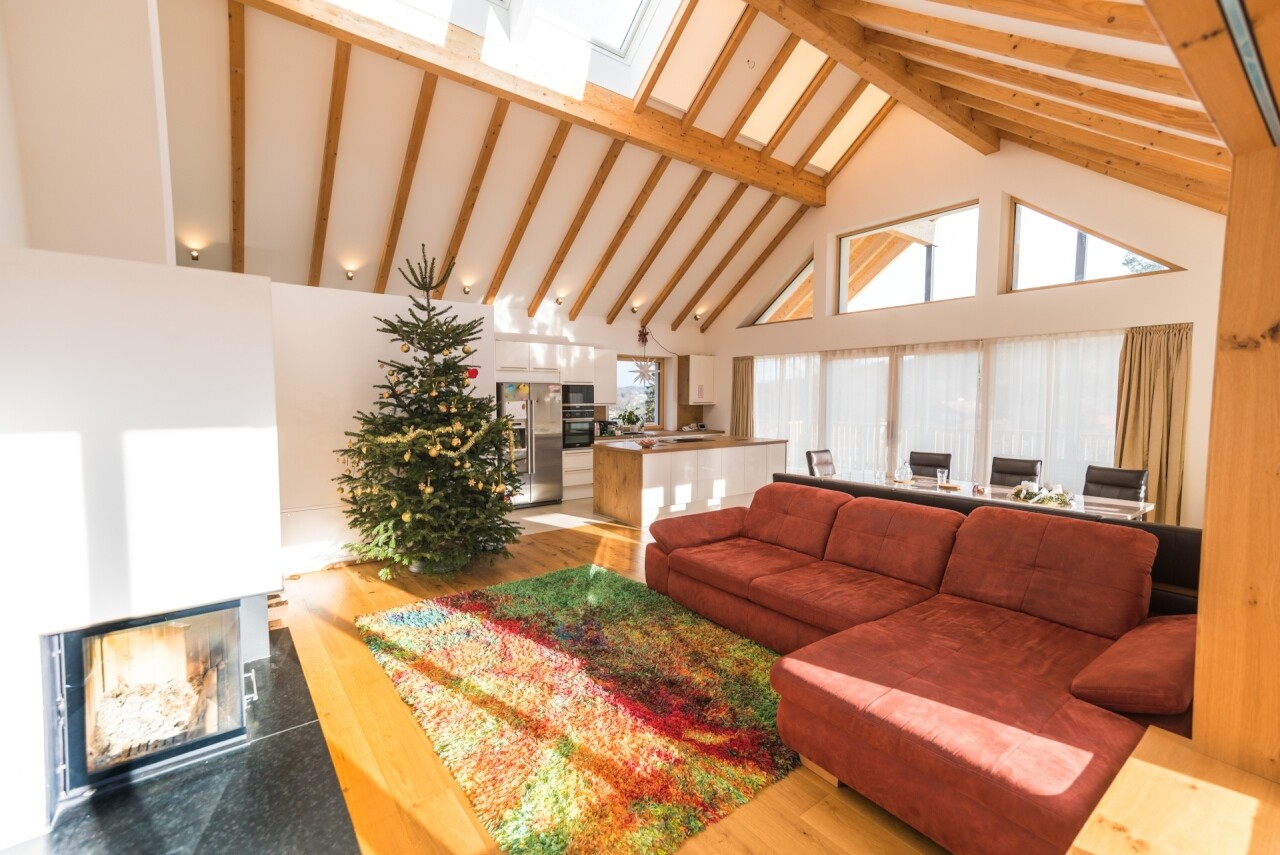 Helles Wohnzimmer mit Holzbalkendecke, roter Ecksofa, bunter Teppich, Weihnachtsbaum und Küche.