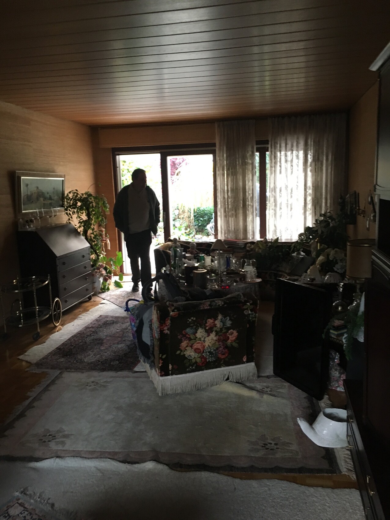 Mann steht im Wohnzimmer an der Tür zum Garten; viele Möbel, Pflanzen und florales Sofa.