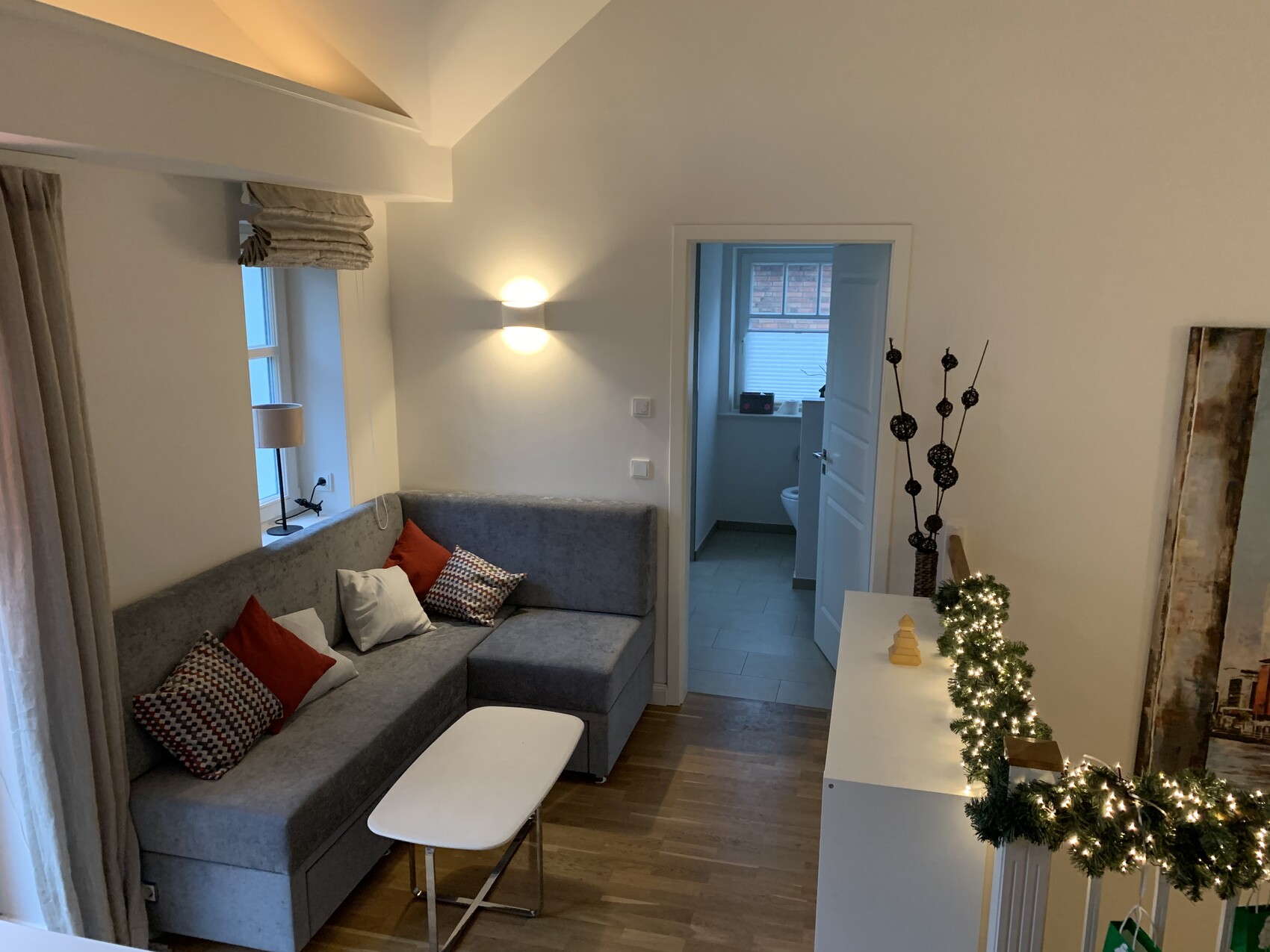 Wohnzimmer mit grauer Eckcouch, roten und gemusterten Kissen, kleinem weißen Tisch und Lichtergirlande.