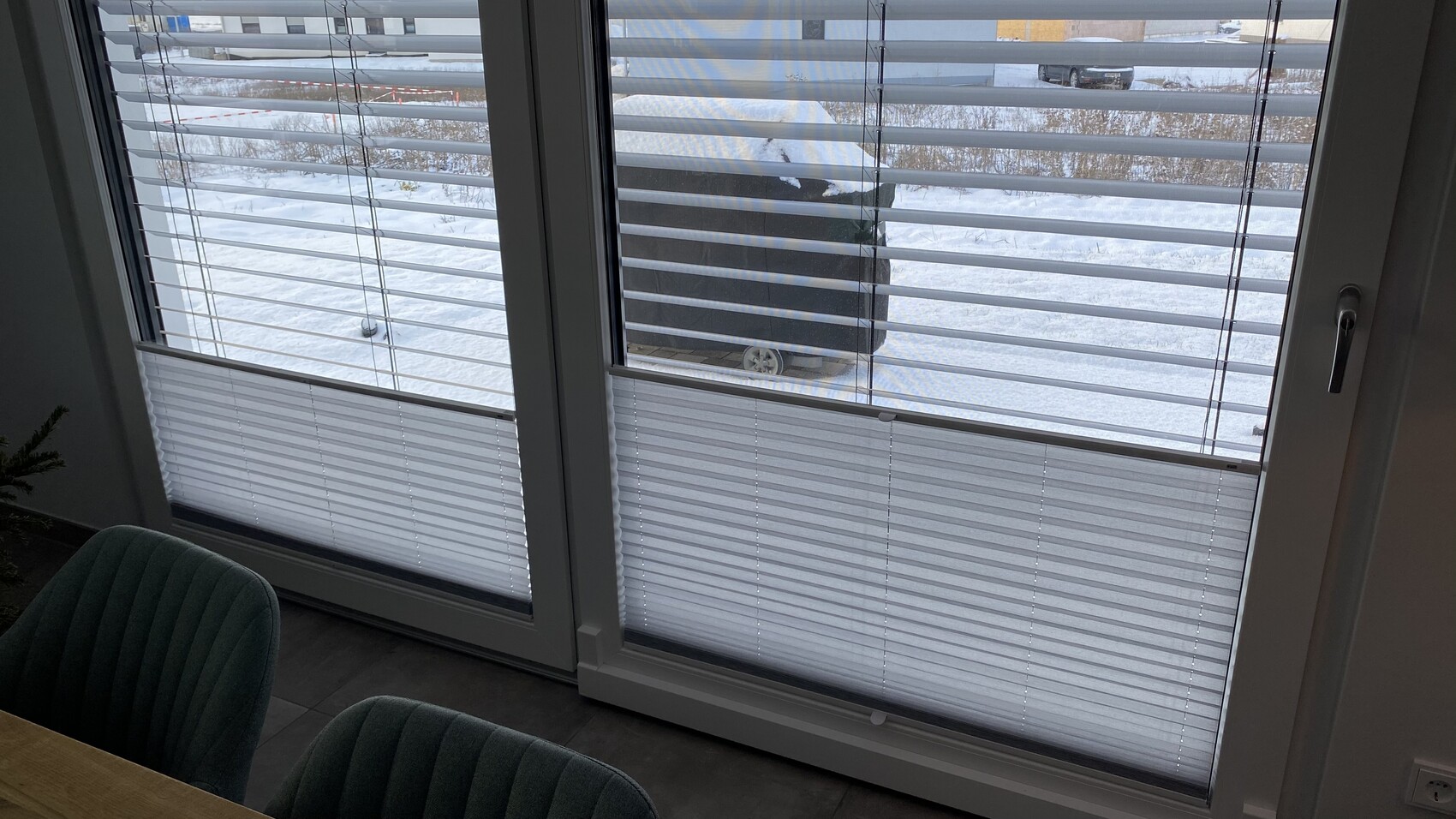 Blick durch Fenster mit horizontalen Jalousien auf verschneiten Außenbereich; Vordergrund grüne Stühle.