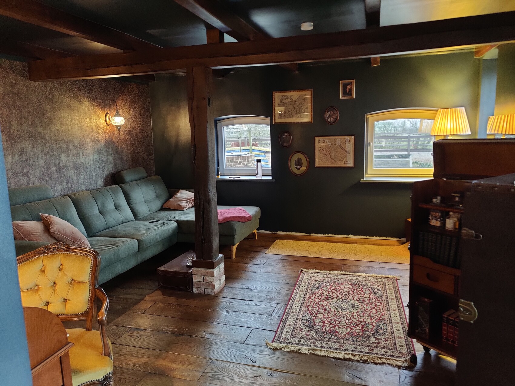 Rustikales Dachboden-Wohnzimmer mit grünem Sofa, Holzboden und Teppich, Blick nach draußen.