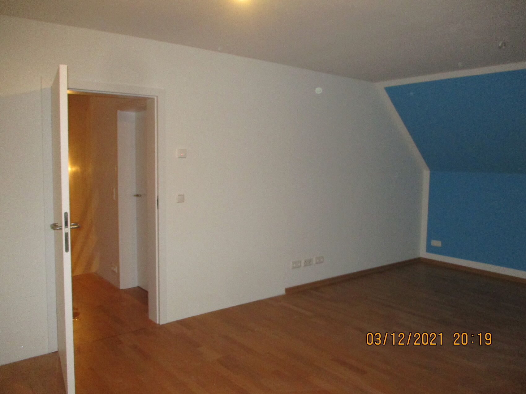Leeres Zimmer mit Holzboden, weiße Wand, blaue Akzentwand rechts, offene Tür zum Flur.