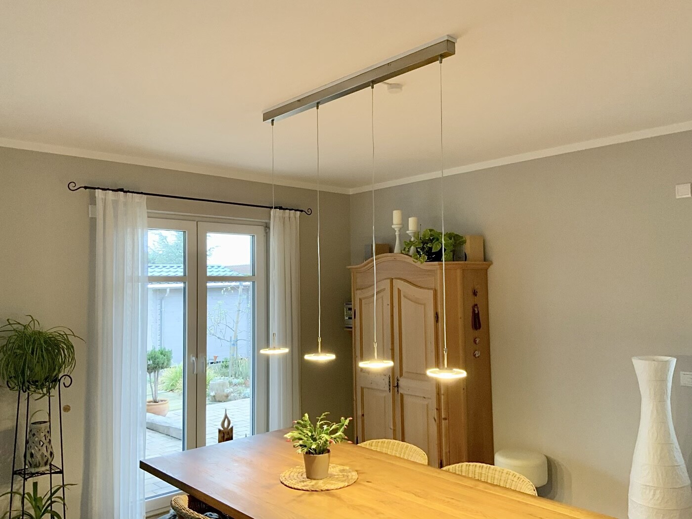 Heller Essbereich: langer Holztisch, vier Pendelleuchten, Holzschrank, Pflanzen, Glastür mit Vorhang.