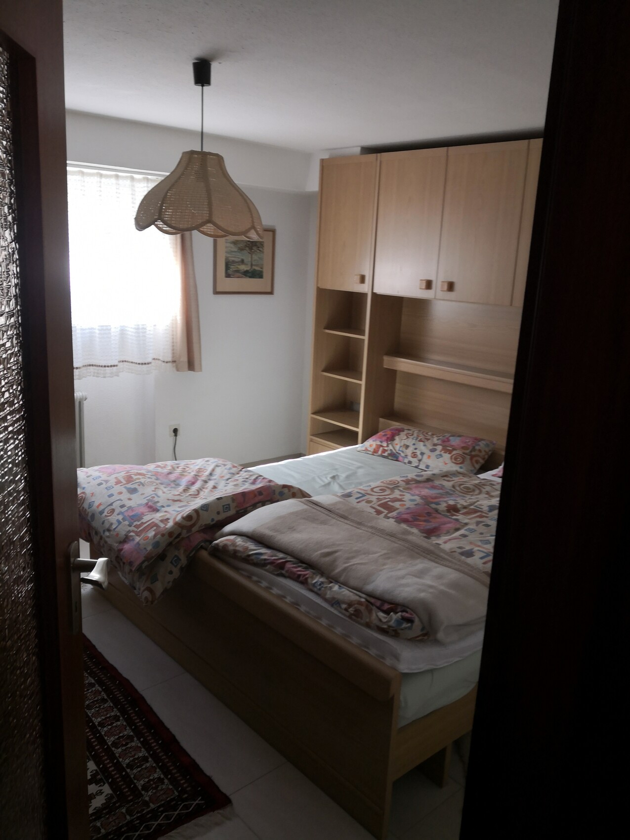 Kleines Schlafzimmer mit Bett, bunt gemusterter Bettwäsche, Holzschrank und Hängeleuchte.