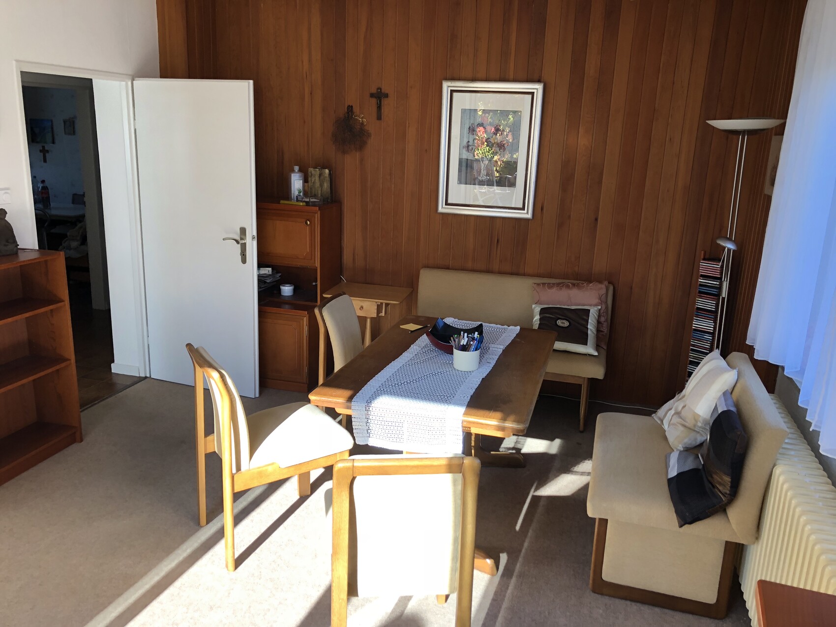 Wohnzimmer mit Holzwand, Esstisch, Stühle, beige Sofa, gerahmtes Bild, Stehlampe und Regal.