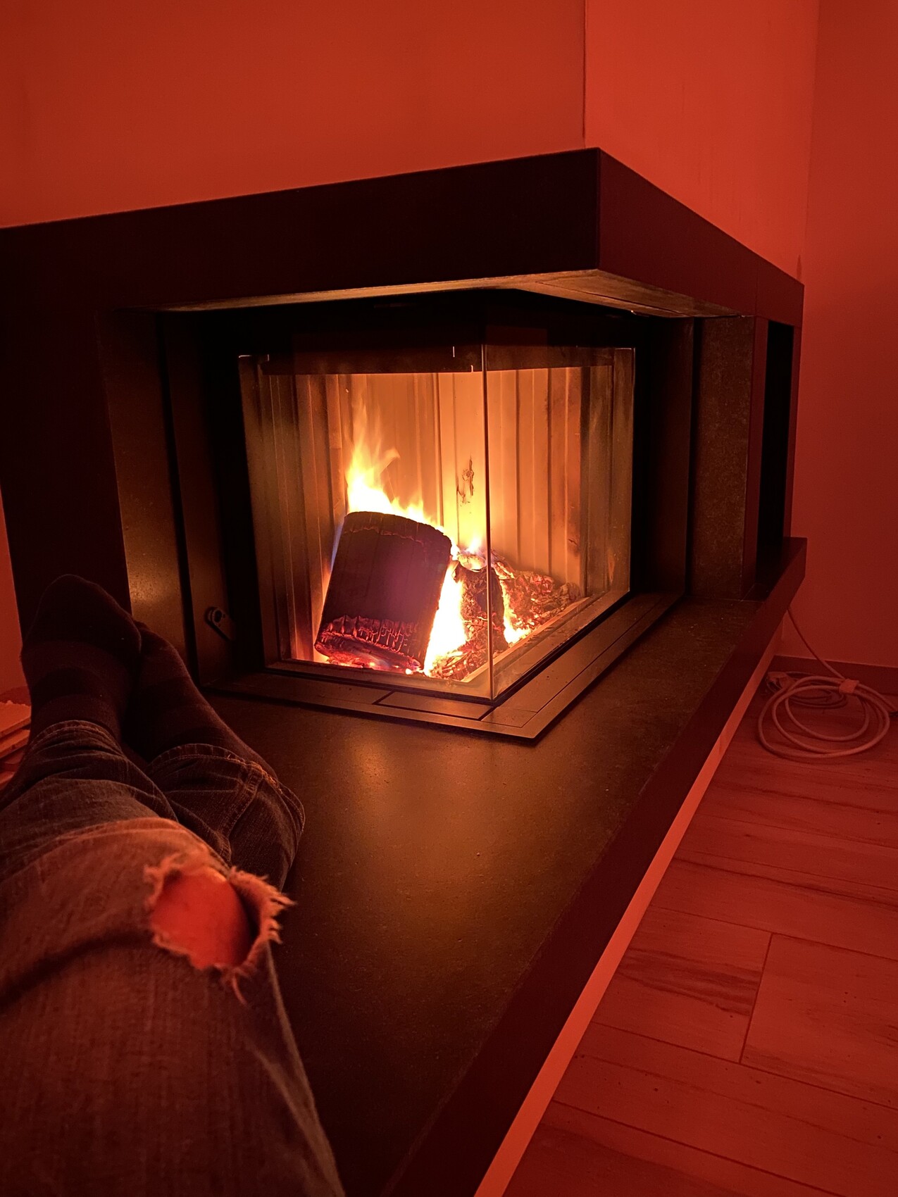 Beine einer Person vor einem Kamin mit brennendem Feuer, rote Lichtstimmung; Jeans mit Loch.