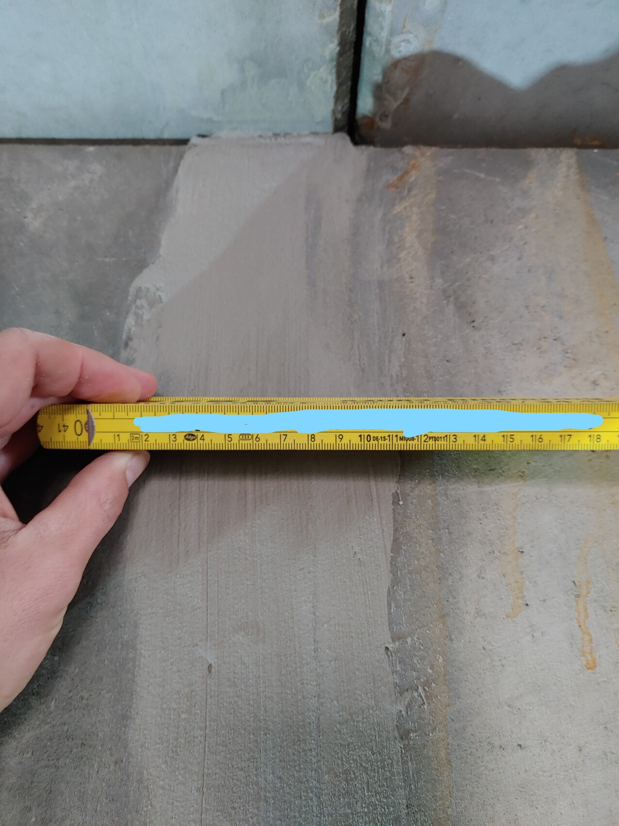 Hand hält ein gelbes Maßband über eine Steinplatte; blauer Streifen markiert die Länge.