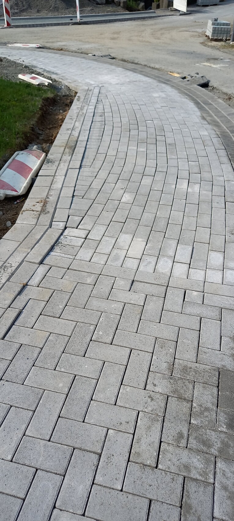 Gepflasterter, kurvenförmiger Gehweg aus grauen Pflastersteinen; Baustelle im Hintergrund.