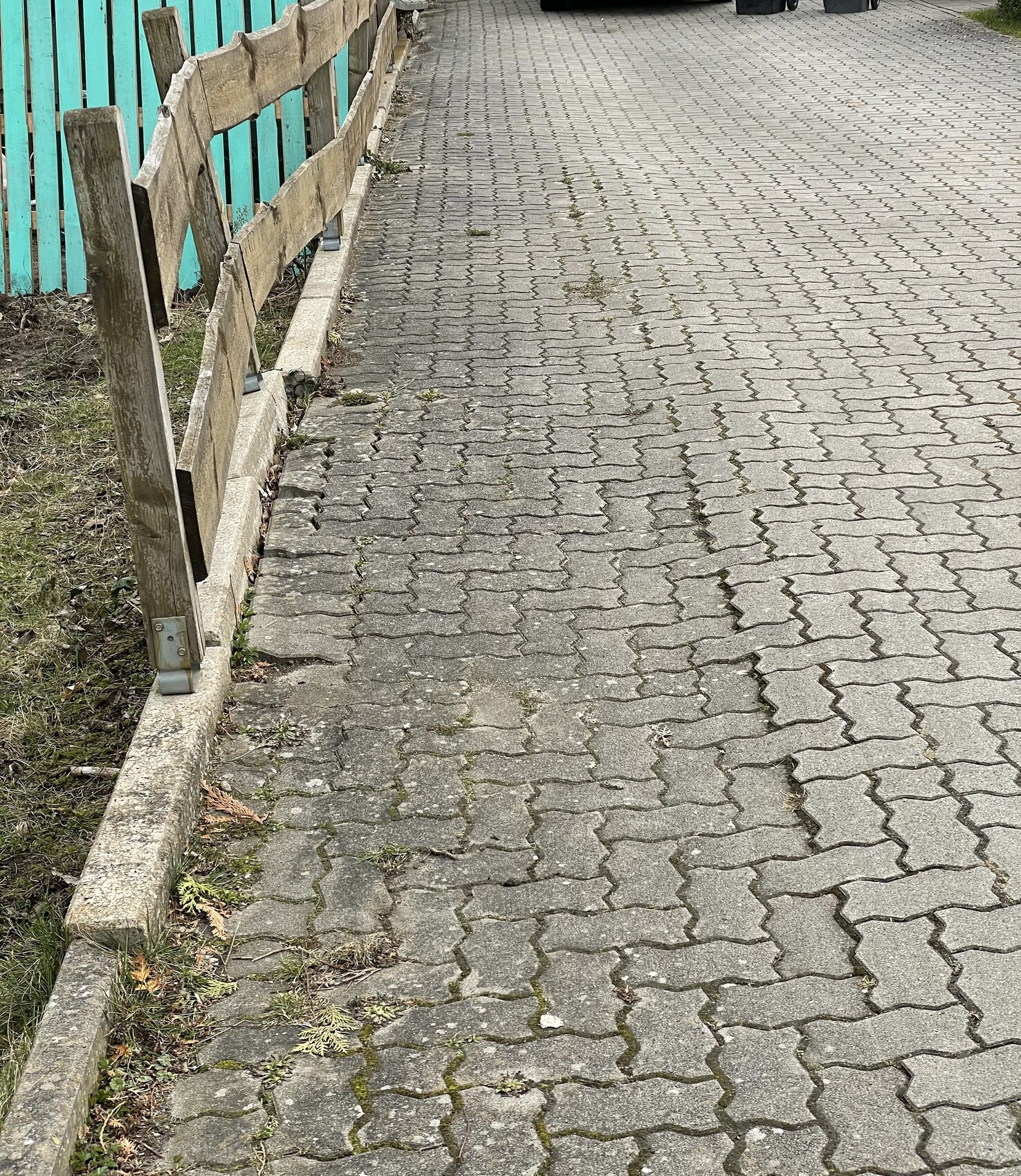 Gepflasterter Gehweg aus Betonpflaster mit Holzlattenzaun links; türkisfarbener Zaun dahinter.
