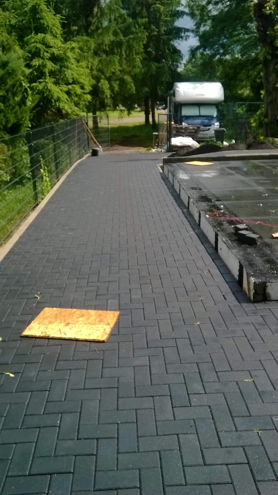 Gepflasterter Pfad im Freien, linker Zaun, Bauarbeiten und Wohnmobil im Hintergrund.