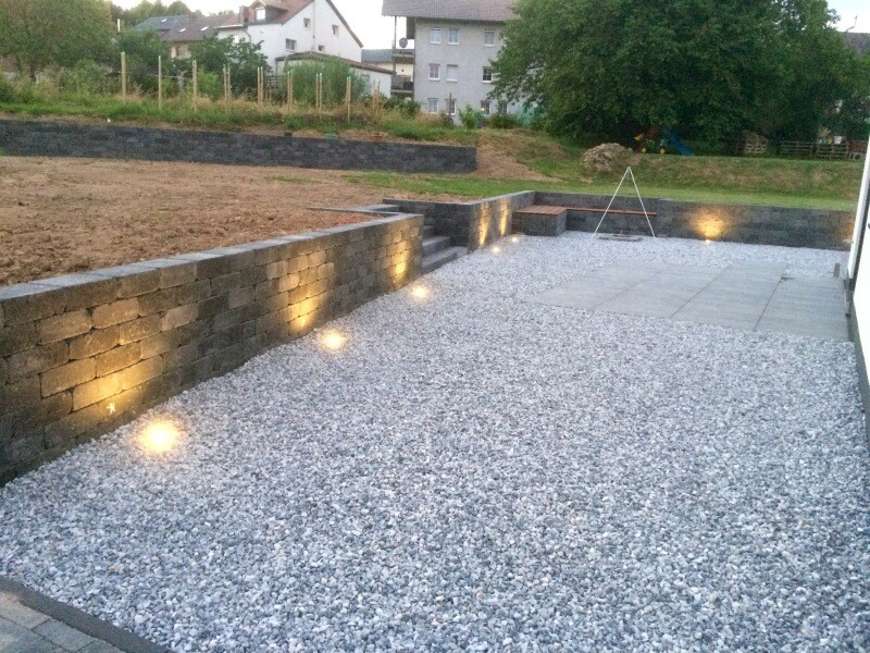 Gartenbereich mit Steinmauer, Kiesboden, Stufen und bodennahe Beleuchtung.