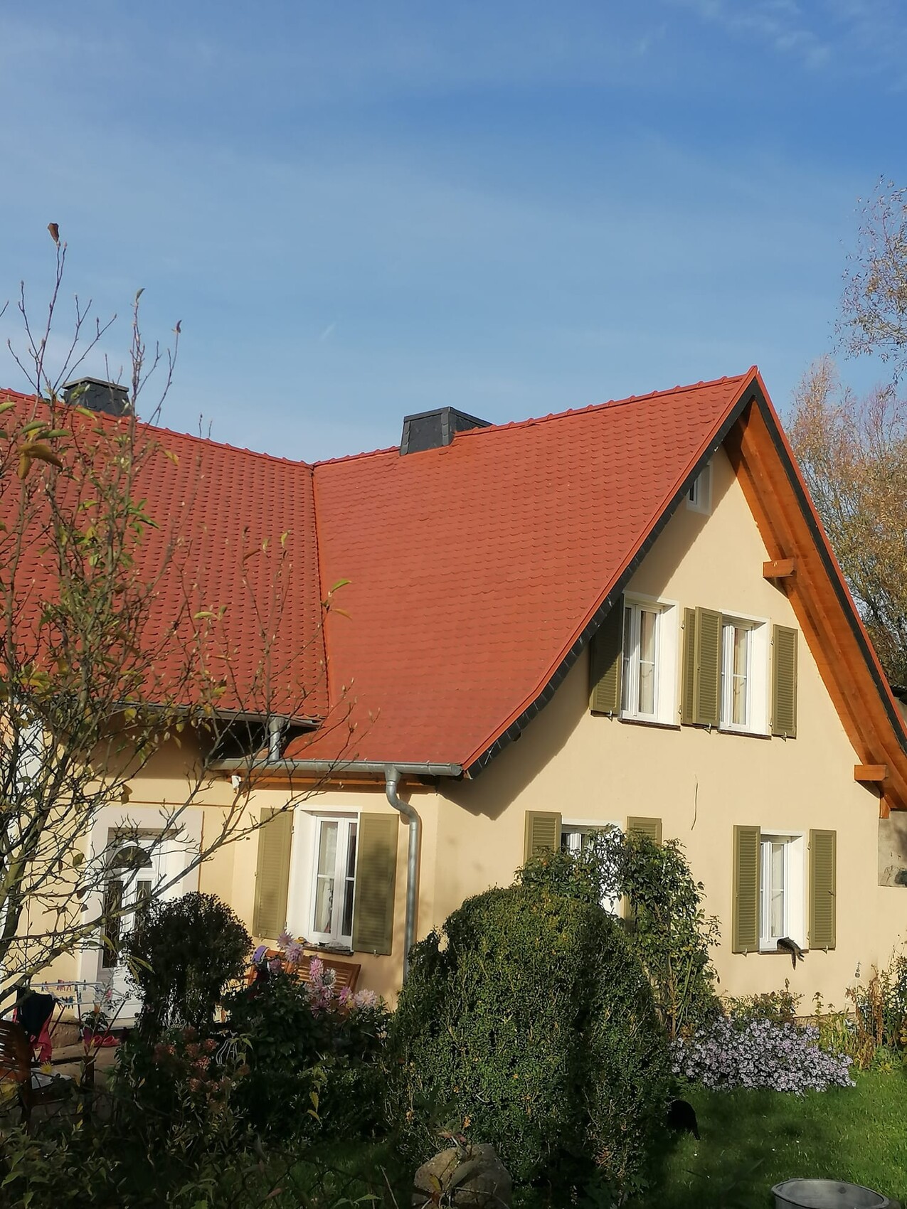 Zweistöckiges Haus mit rotem Ziegeldach, beigefarbener Fassade, grünen Fensterläden, Garten.