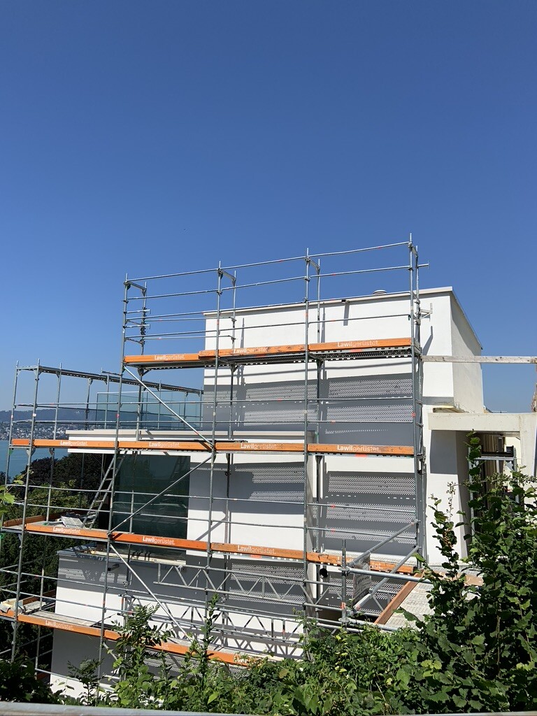 Weißes Gebäude mit Gerüst aus Stahl und orange Bretter, blauer Himmel und Grün im Vordergrund