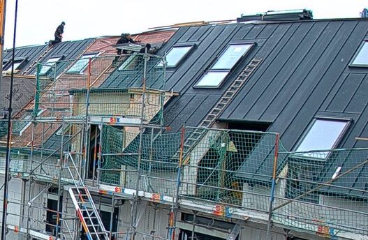 Mehrstöckiges Gebäude mit dunklem Metall-Dach, Bauarbeiter arbeiten auf dem Gerüst.