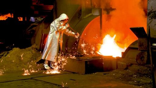 Person in Schutzanzug arbeitet neben heißem Metall in Gießerei; Funken fliegen.