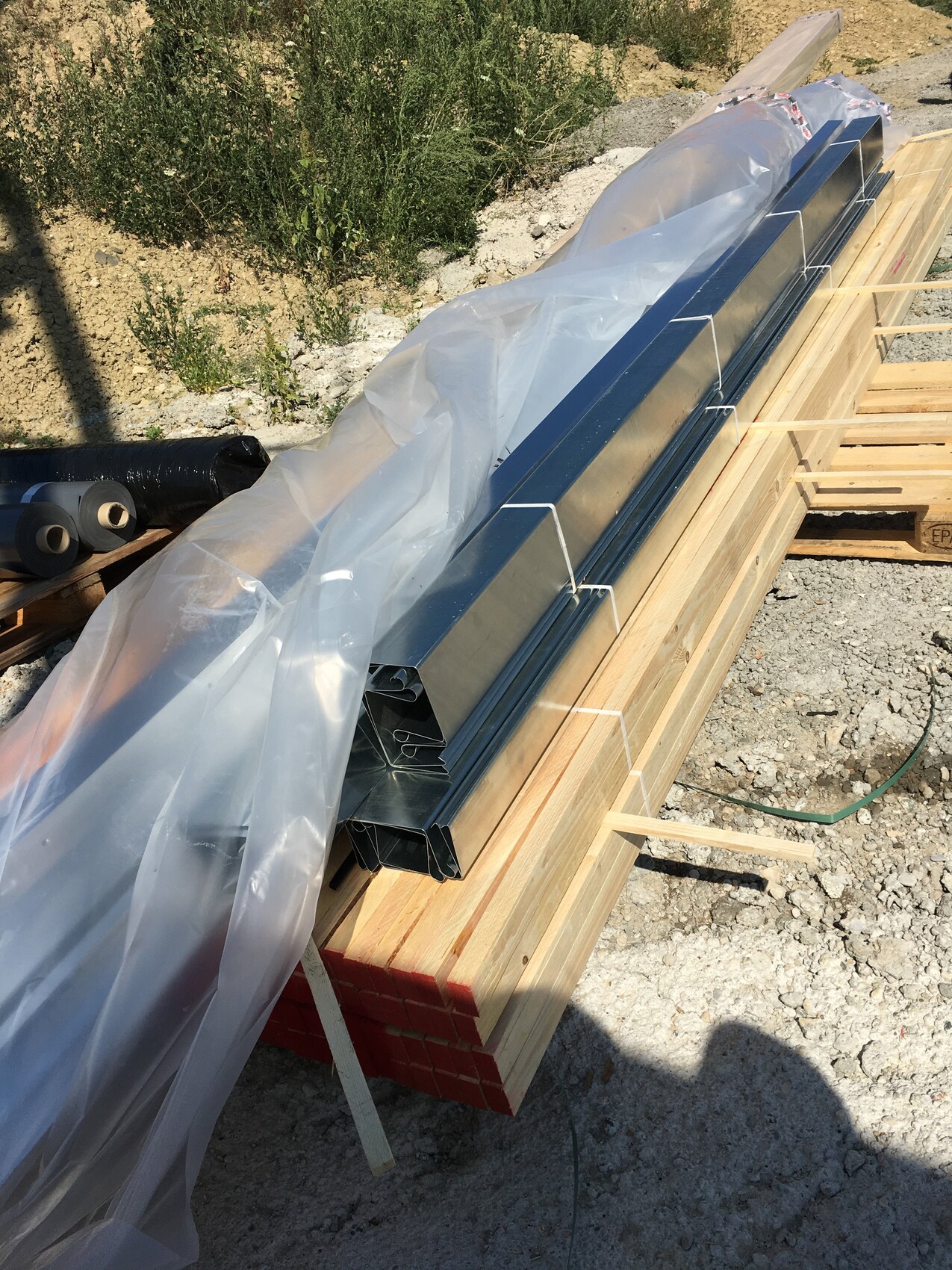 Gestapelte verzinkte Metallprofile auf Holzbalken am Bauplatz, teilweise in Plastik eingewickelt.