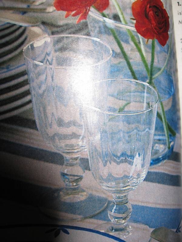 Zwei klare Weingläser stehen auf einem Tisch, im Hintergrund rote Rosen in einer Vase.