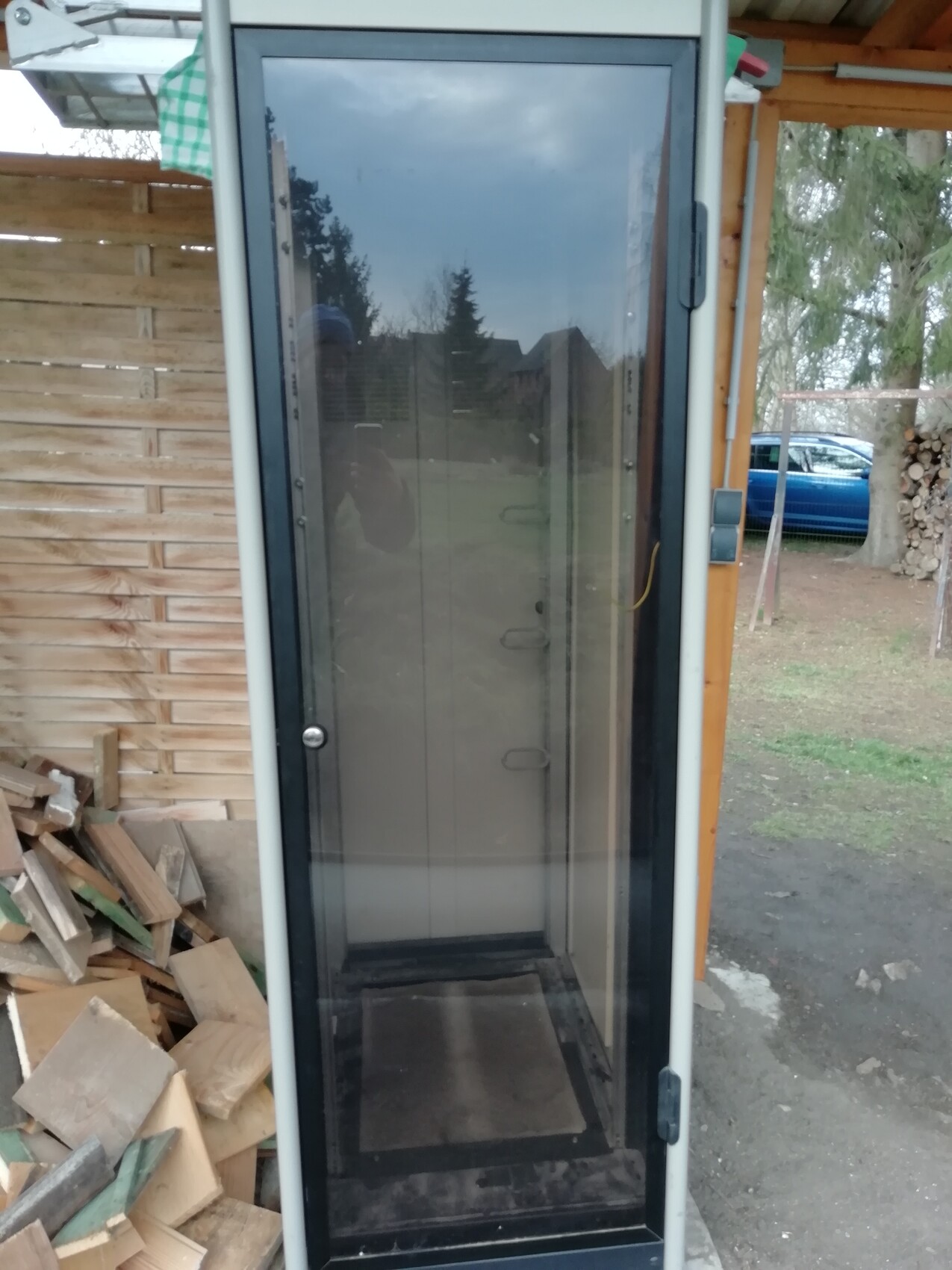 Freistehende Glasvitrine im Außenbereich neben Holzstapeln; Spiegelung des Fotografen