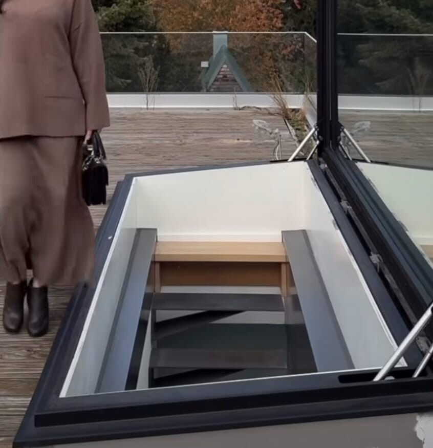 Person in beige Mantel steht auf einer Terrasse neben einem offenen Glasboden mit Treppe nach unten.