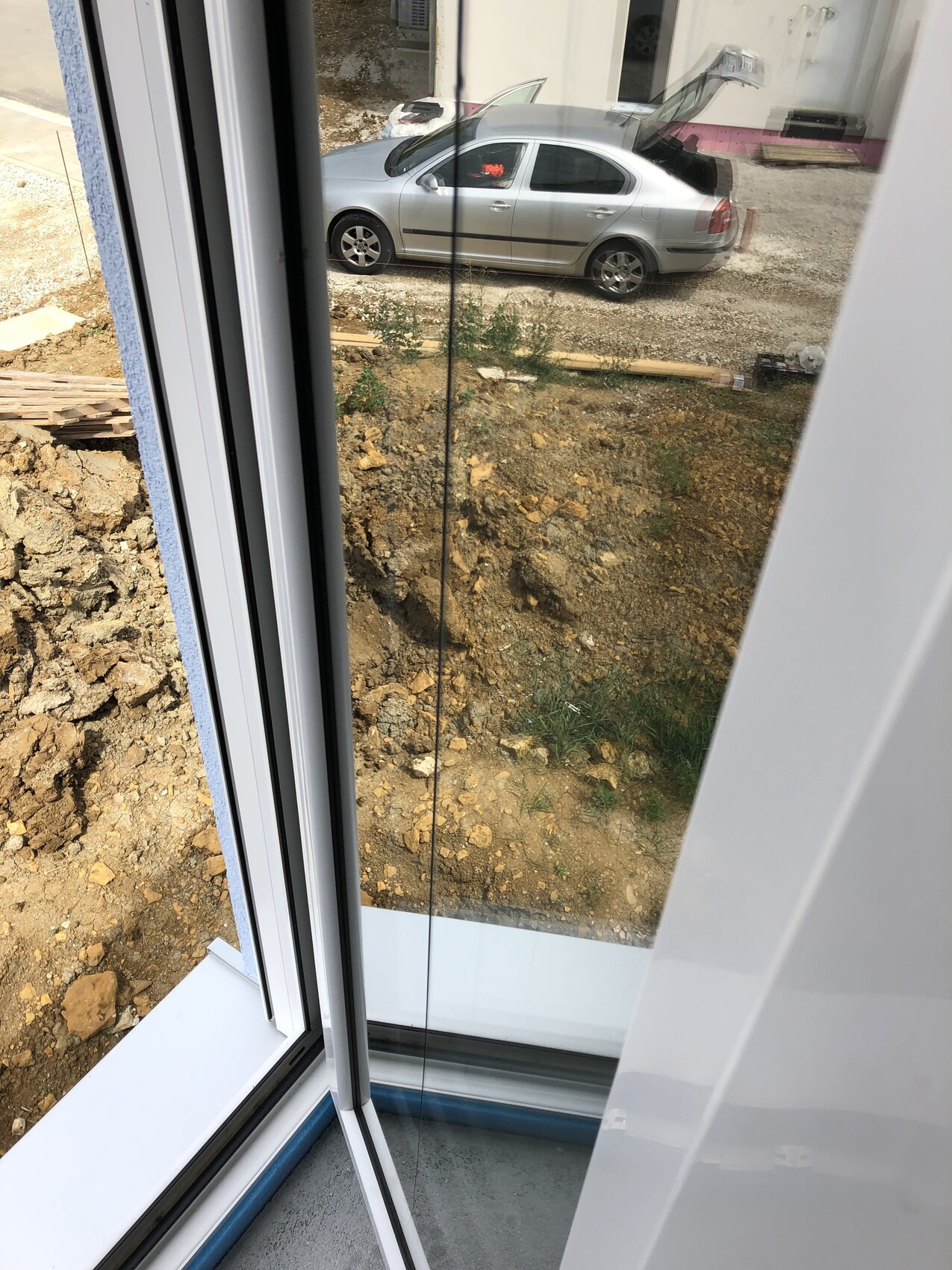 Blick durch geöffnete Glasschiebetür auf silbernen Pkw mit offener Heckklappe vor Baugelände.