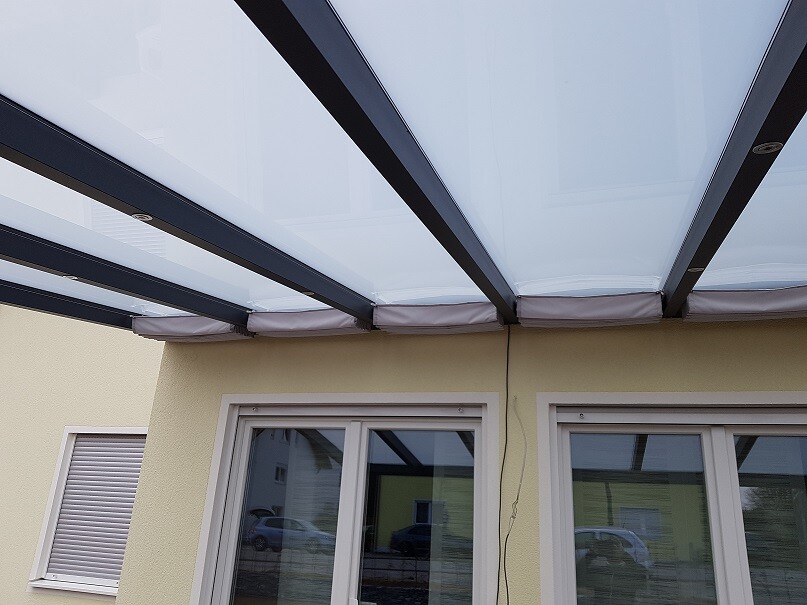 Glasdach mit schwarzen Stahlträgern über Terrassentüren vor gelber Hauswand.