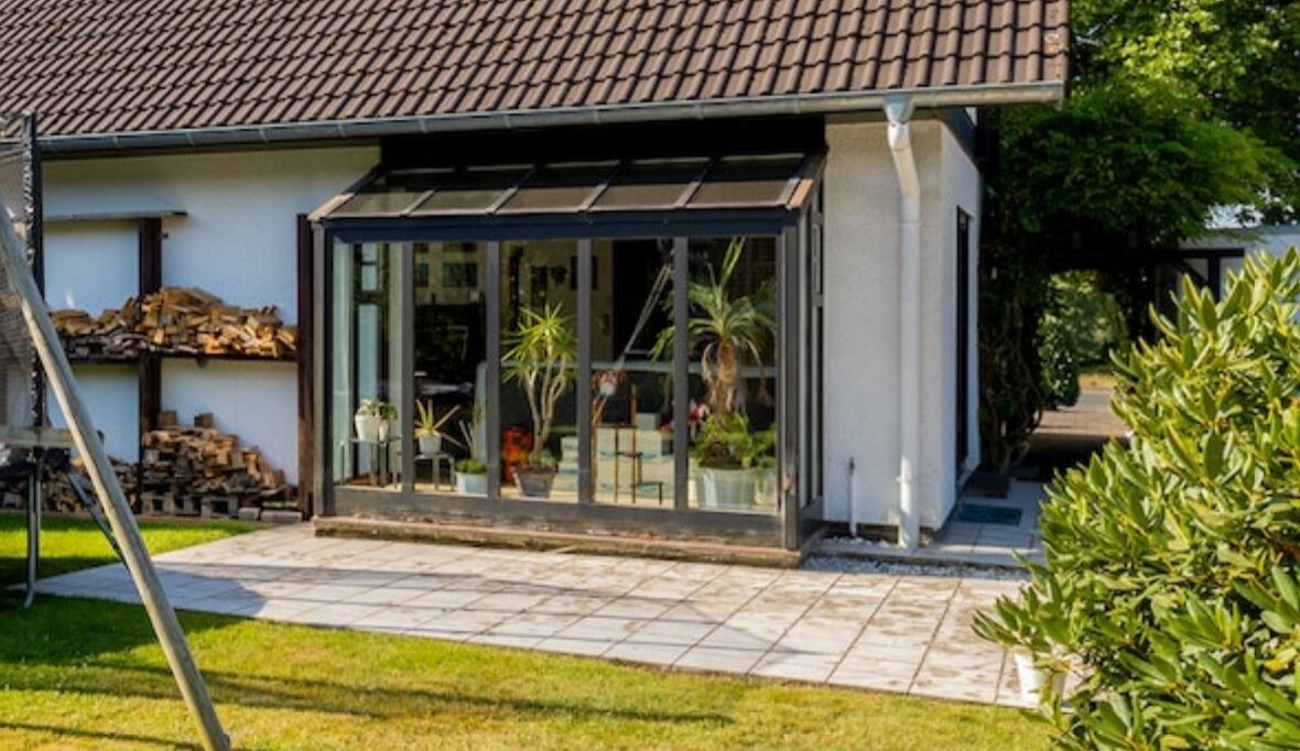 Glas-Wintergarten am Haus mit Topfpflanzen, links Holzstapel, gepflasterte Terrasse.
