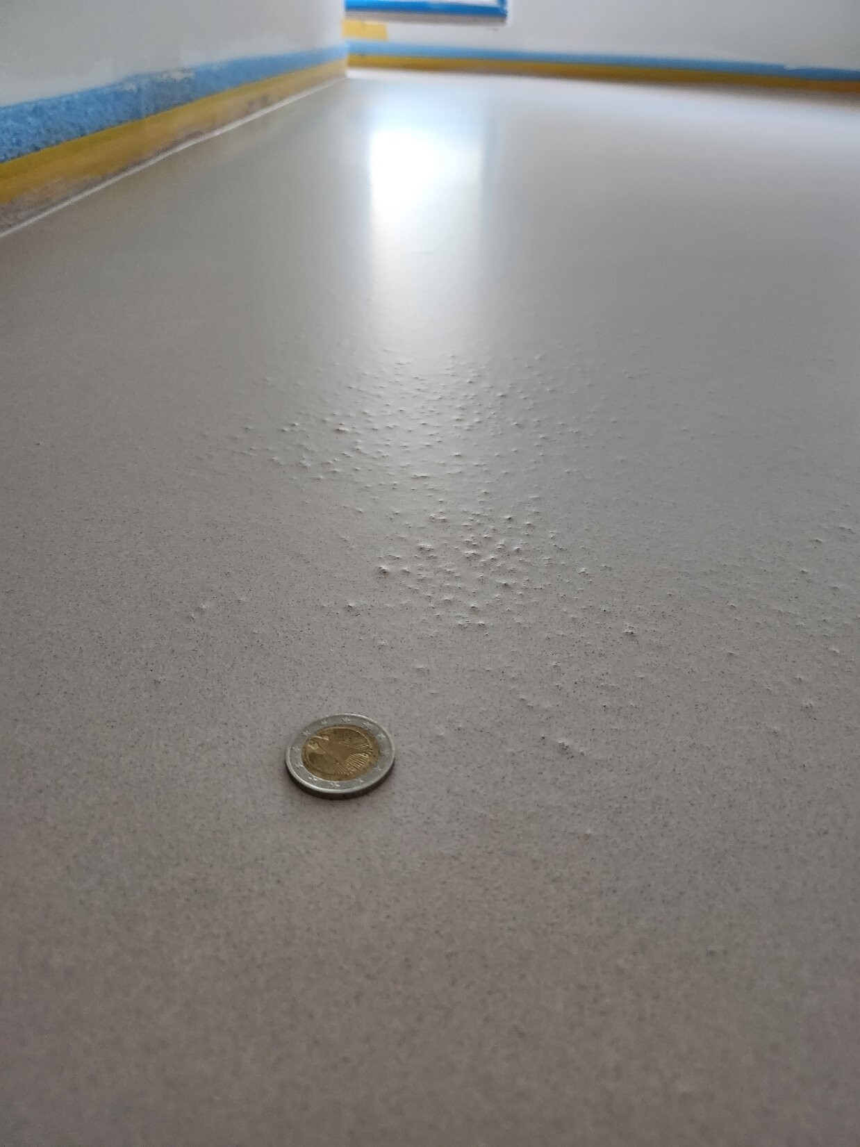 Nahaufnahme einer Euro-Münze auf glattem grauen Boden in Flur, blau-gelber Wandrand.