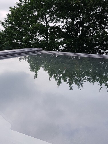 Spiegelndes Autodach mit Baumreflexionen und Wolken am Himmel.