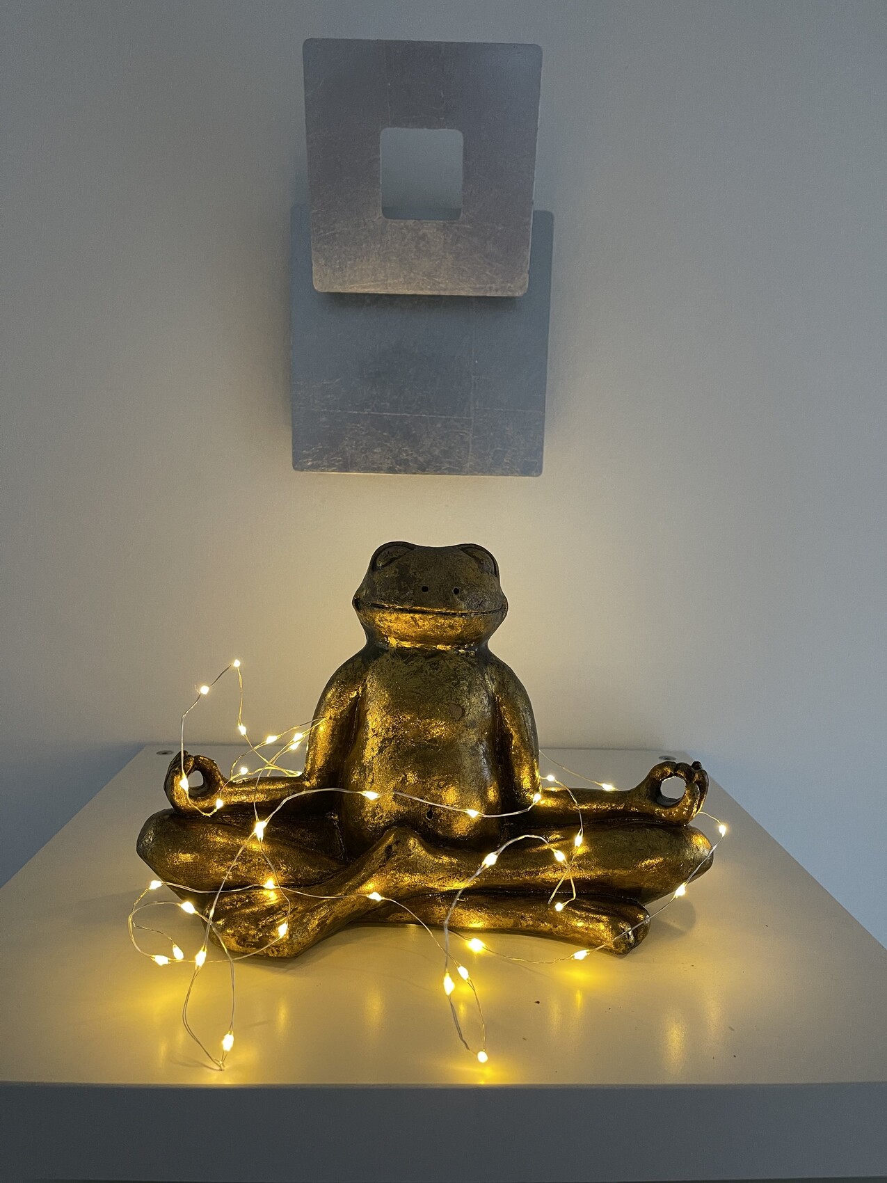 Goldene Froschfigur in Meditationshaltung mit Lichterkette auf weißem Regal.