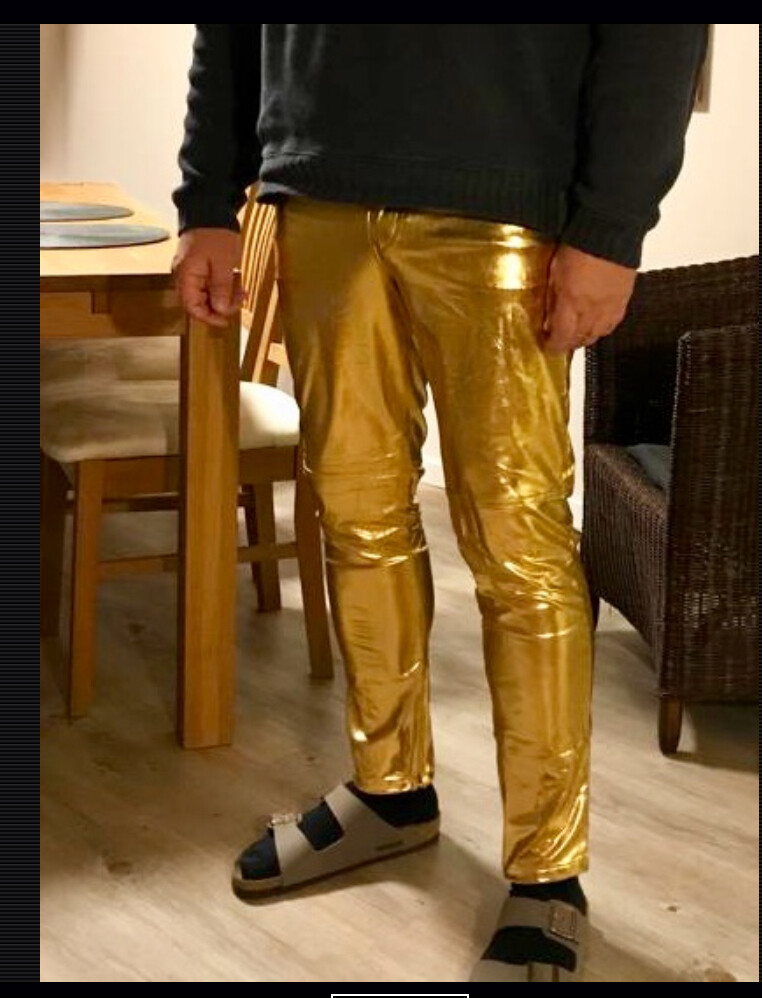 Person steht drinnen, trägt eine glänzende goldene Hose, Sandalen mit Socken, schwarzer Pullover.