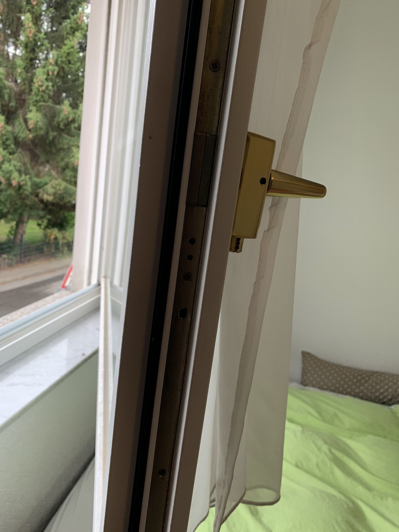 Offener Fensterflügel mit goldfarbenem Griff; Blick nach außen auf Bäume, Bett im Zimmer.