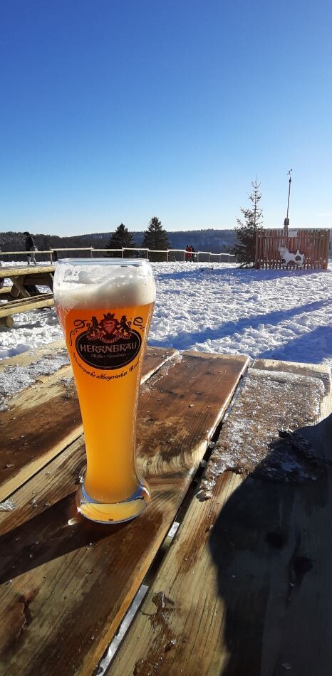 Helles Bierglas mit Schaum auf Holzbank im Schnee, Winterlandschaft und blauer Himmel.