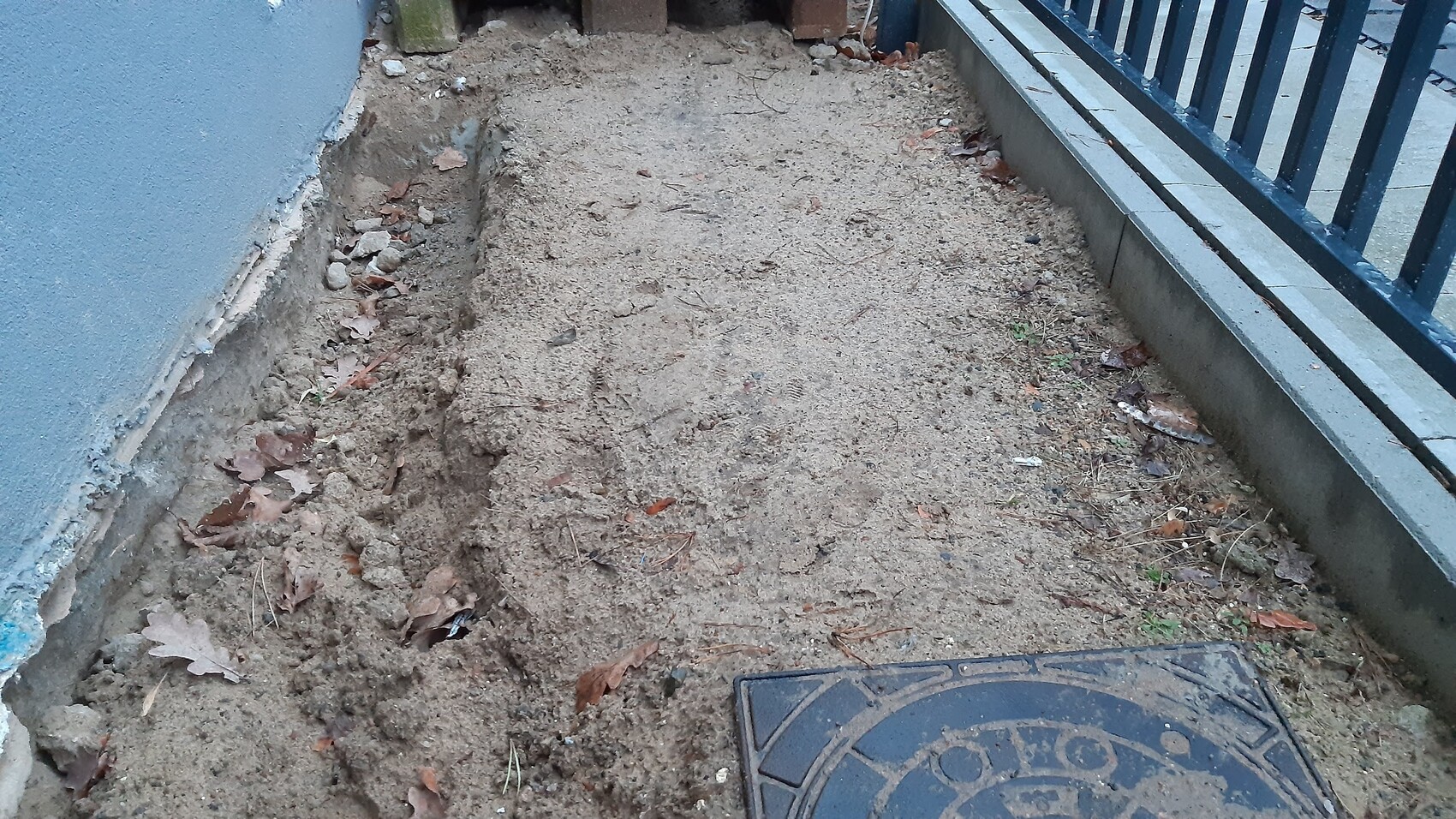Schmaler Erdstreifen neben blauer Wand mit Geländer, Sandboden und Blättern.