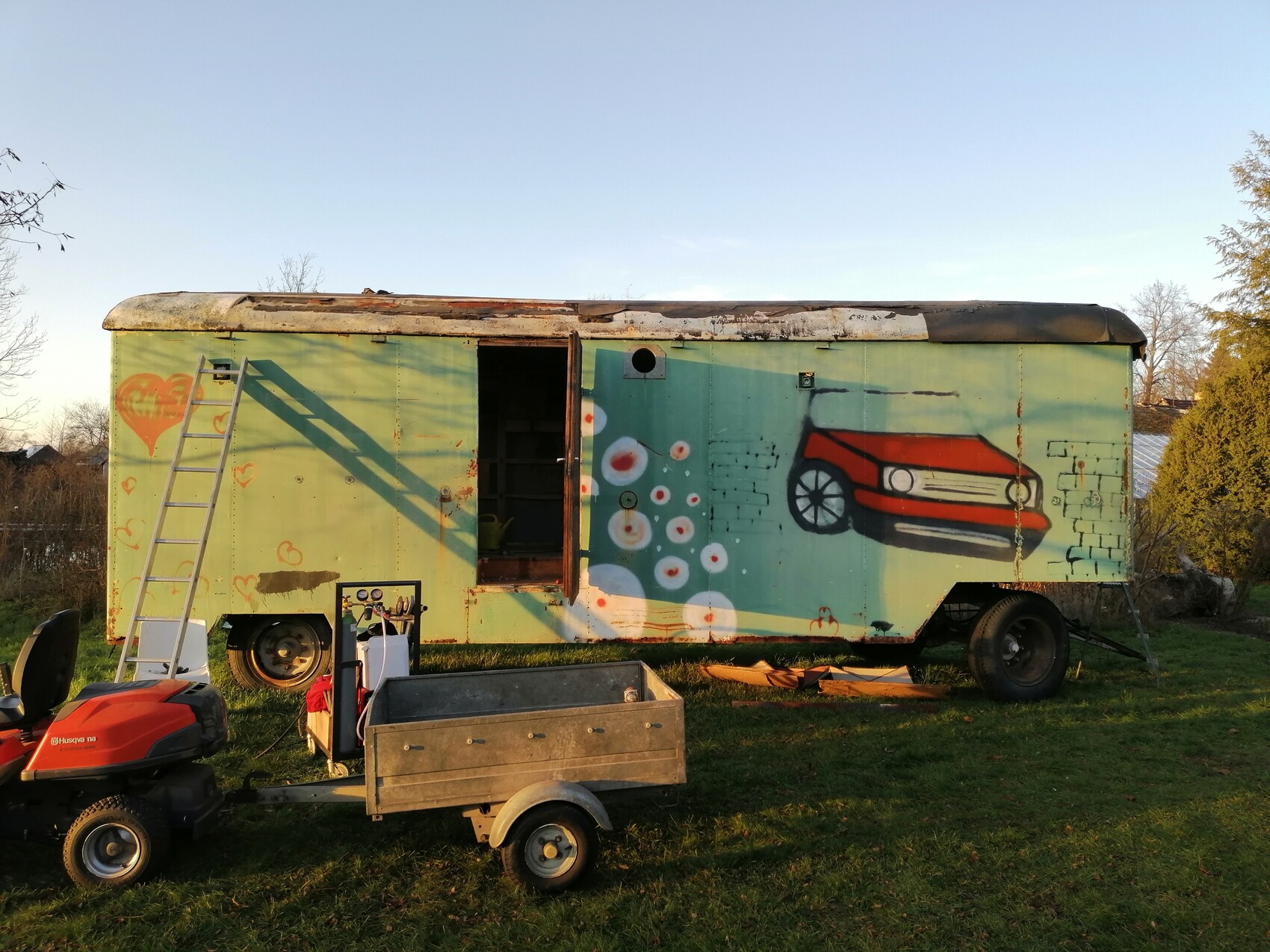 Graffiti-Trailer auf grünem Gartenplatz mit Leiter und Rasenmäher im Freien
