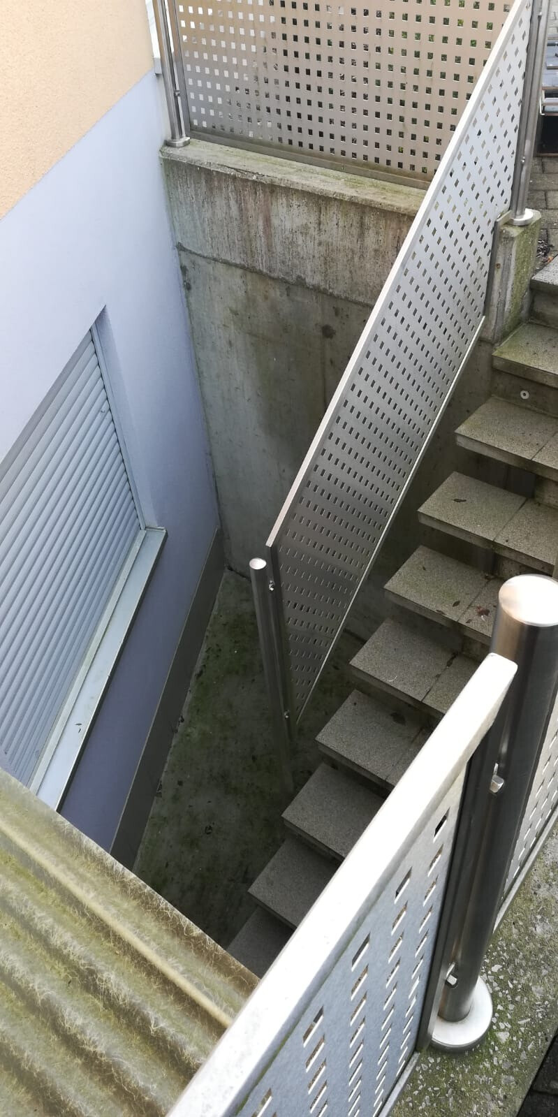Blick von oben auf eine Außentreppe mit Stufen, Lochblech-Metallgeländer und Betonwand.