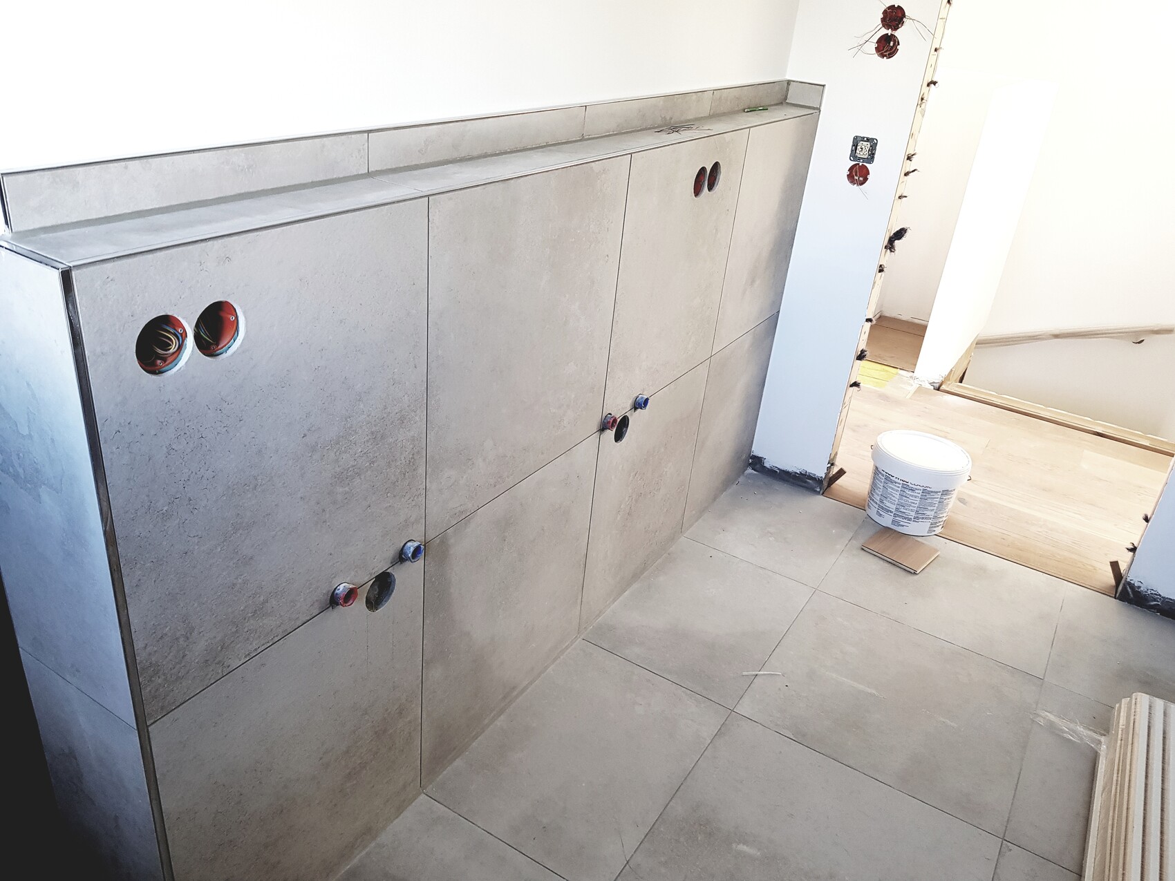 Badezimmerunterbau aus grauen Fliesen; offene Rohranschlüsse, Eimer auf dem Boden.