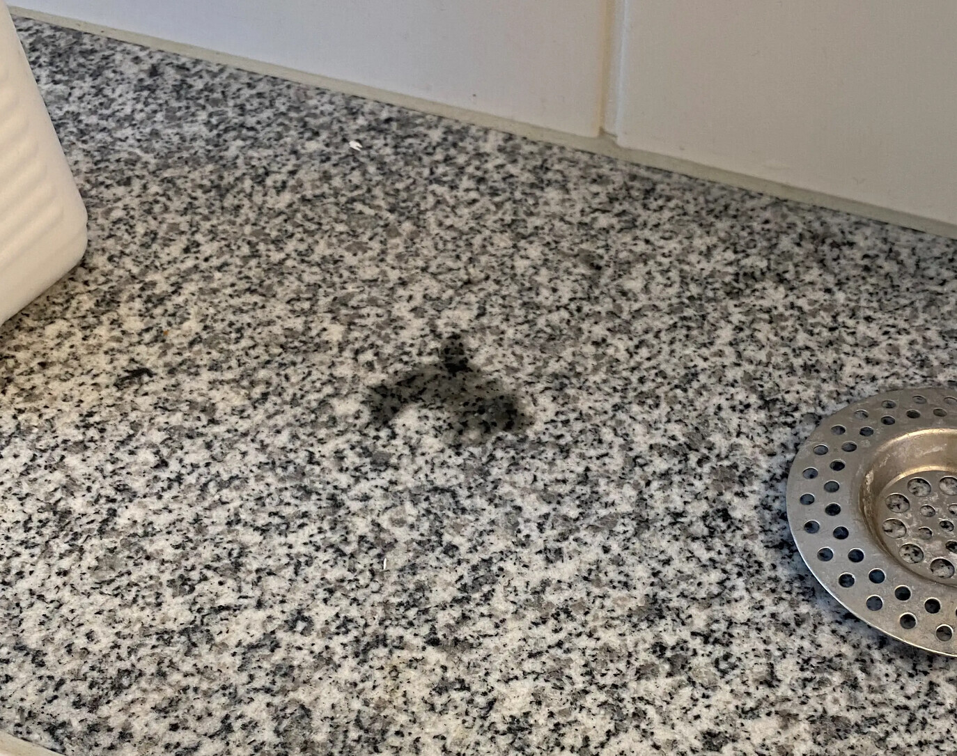 Graue Granit-Arbeitsplatte mit runder Abflussabdeckung rechts; dunkler Fleck in der Mitte.