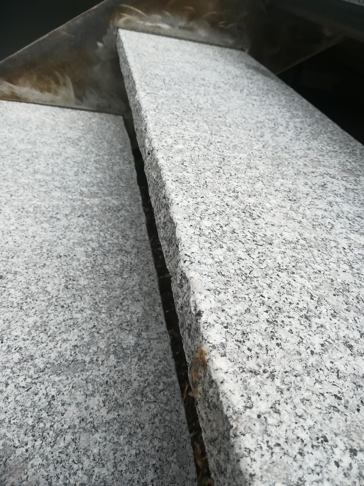 Nahaufnahme grauer Granitplatten mit Spalt dazwischen; rostige Metallkante oben.